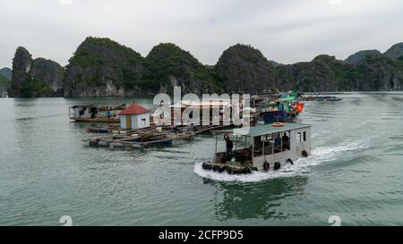 Cat Ba, Vietnam - 19 novembre 2019 : bateau de pêche avec coquillages passe village flottant, près de Ha long Bay, karst de montagne, ciel couvert. Banque D'Images