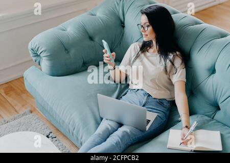 Photo d'une femme indépendante occupée dans des vêtements décontractés axés sur l'affichage de smartphone, vérifie l'information sur Internet, note quelque chose dans le journal, utilise moderne Banque D'Images