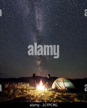 Couple romantique de touristes se reposant au camping de nuit d'été dans les montagnes. Vue arrière de l'homme et de la femme assis sur des chaises à côté du feu de camp et de la tente, en tenant les mains sous le ciel étoilé plein d'étoiles et de voie lactée. Banque D'Images