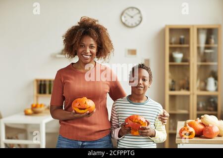 Portrait horizontal moyen de la jeune femme adulte et de sa préadolescence fils debout ensemble dans la salle de séjour tenant des citrouilles qu'ils ont sculptées sourire à l'appareil photo Banque D'Images