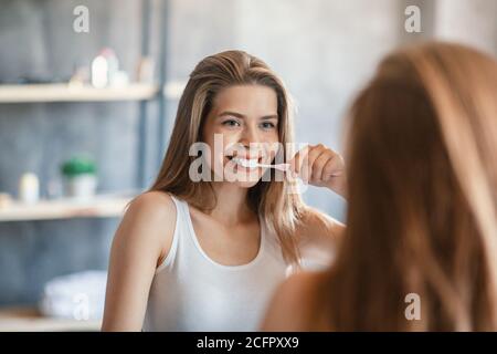 Jolie jeune femme avec un beau sourire se brossant les dents face du miroir dans la salle de bains Banque D'Images