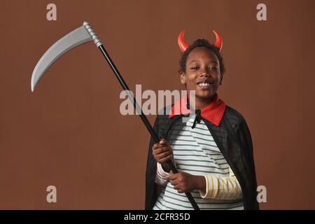 Studio portrait d'un garçon joyeux de la préadolescence afro-américaine portant des cornes rouges sur la tête et le manteau costume tenant scythe regardant l'appareil photo sourire, fond marron Banque D'Images