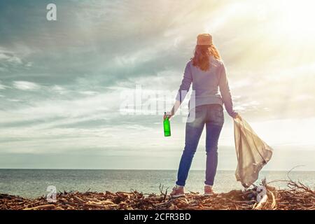 La femme bénévole se tient sur le rivage de la mer. Elle tient un sac à ordures dans une main et une bouteille sale dans l'autre. Nettoyage de la plage après un St Banque D'Images