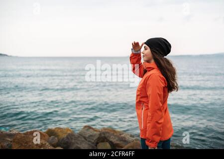 Une femme brune aux cheveux longs portant une casquette et une veste orange se tient sur le rivage et regarde la distance. En arrière-plan la mer et le Banque D'Images