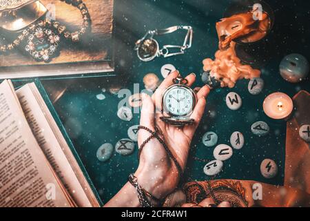 Astrologie et ésotéricisme. La main femelle de la sorcière tient une montre sur une chaîne. En arrière-plan, les vieux livres, les runes qui disent de la fortune, les bougies et je Banque D'Images