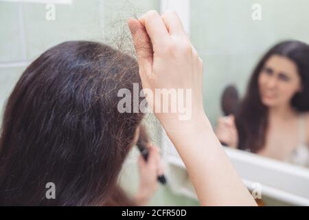 Une femme brune tire une bande de cheveux de sa tête. Vue arrière. En arrière-plan, une réflexion dans le miroir. Le concept de la perte de cheveux. Banque D'Images