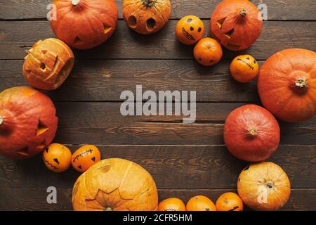 Plan de coupage plat de dessus en bas de citrouilles mûres d'orange sculptées et de mandarines avec des visages dessinés pour Halloween allongé sur une table en bois marron dans un cercle, espace de copie Banque D'Images