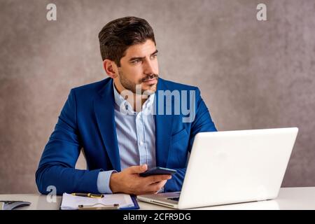 Photo courte d'un homme d'affaires stressé tenant son téléphone portable dans la main tout en étant assis derrière son ordinateur portable et en regardant avec soin. Bureau à domicile. Banque D'Images