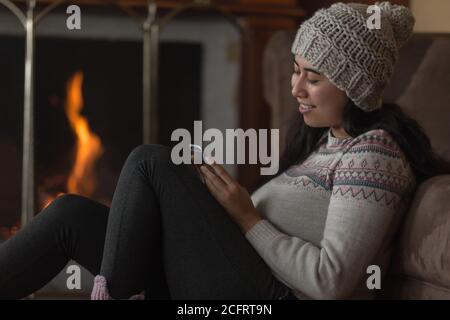 Une jeune femme latine souriante assise sur le côté avec un téléphone portable tenant sa main et touchant sa tête, avec une jambe tendue, porte un chapeau en laine Banque D'Images