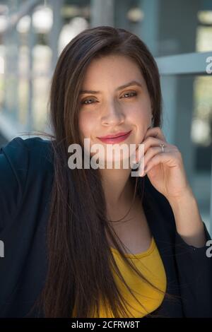 Visage d'une jeune femme latine à la peau blanche avec des cheveux longs et droits, des yeux légers et un maquillage souriant avec une main sur sa joue, regardant droit devant Banque D'Images