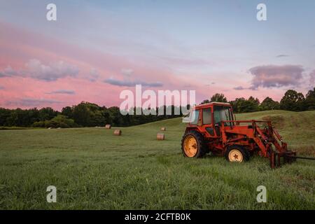 Le tracteur rouge et les balles de foin fraîchement laminées reposent sur le roulement colline avec un paysage de nuages spectaculaire au lever du soleil créant une campagne rustique scène Banque D'Images