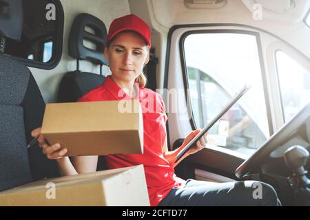 femme employée du service de livraison en uniforme rouge, assise dans une camionnette et travailler avec des packages Banque D'Images