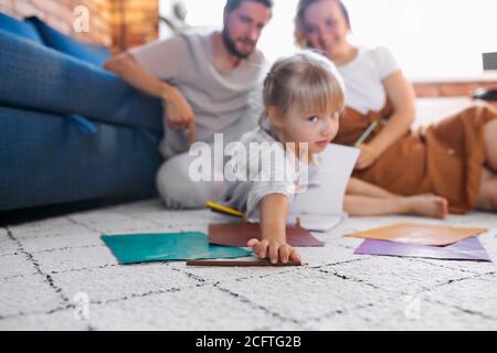 les parents heureux détendus s'assoient avec leur fille sur le sol, la petite fille caucasienne s'assoit à dessiner, avec des papiers à côté d'elle. à la maison Banque D'Images