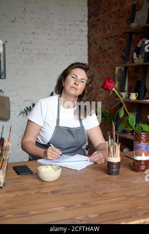 Femme artisanale écrivant dans un carnet, planifiant la journée dans un journal. Belle femme artisan assis à l'atelier dans la chaise et de penser à travailler sur des projets, regardant la caméra Banque D'Images