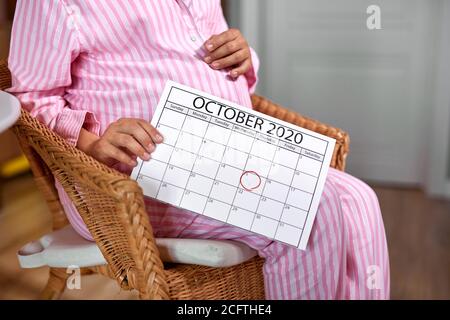 femme enceinte tenir le calendrier dans les mains, attendez-vous à bébé. en comptant les jours, elle donnera naissance en octobre. asseyez-vous sur la chaise à la maison portant un pyjama Banque D'Images