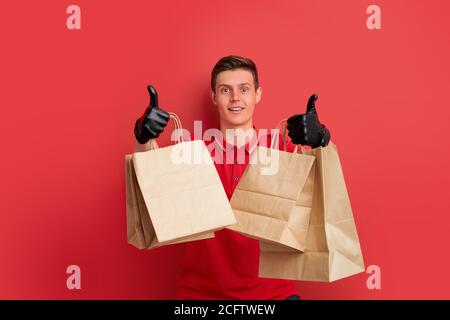 un jeune homme de livraison caucasien, en uniforme de t-shirt rouge et en gants de protection, tient un paquet de papier d'artisanat avec des aliments isolés sur fond rouge, Banque D'Images