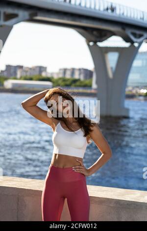 S'adapte jeune femme athlétique qui étire le corps sur un remblai. Fitness femelle dans des leggings roses faire l'entraînement d'échauffement pour les muscles de cou en extérieur. Banque D'Images