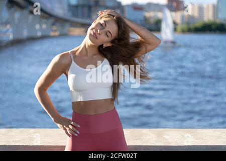 S'adapte jeune femme athlétique qui étire le corps sur un remblai. Fitness femelle dans des leggings roses faire l'entraînement d'échauffement pour les muscles de cou en extérieur. Banque D'Images