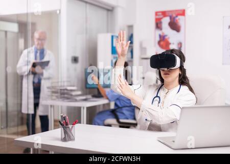 Médecin portant des lunettes de réalité virtuelle de haute technologie dans le bureau de l'hôpital et un manteau blanc avec stéthoscope. Un médecin expérimenté écrit le diagnostic du patient sur le presse-papiers et l'infirmière tient une image radiologique homme malade. Banque D'Images