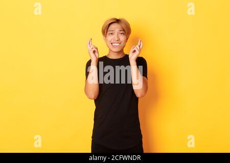 Portrait de l'asiatique nerveux de gars croiser les doigts bonne chance, faisant le souhait, priant sur fond jaune Banque D'Images