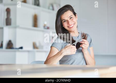 Bonne jeune femme utilisant des ciseaux à la maison tout en la coupant cheveux longs par elle-même Banque D'Images