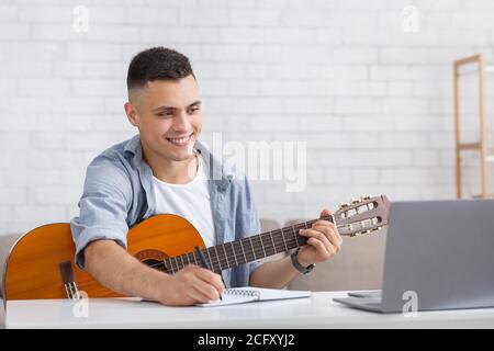 Jeune homme souriant regardant la leçon en ligne, jouant de la guitare et prend des notes Banque D'Images