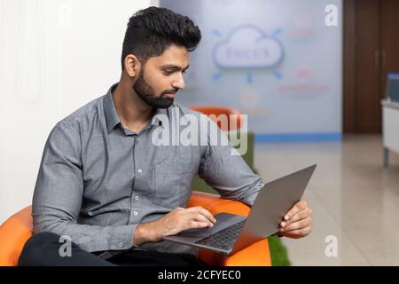 Portrait d'un jeune homme d'affaires indien prospère assis sur un canapé et travaillant sur un ordinateur portable, environnement d'entreprise. Banque D'Images