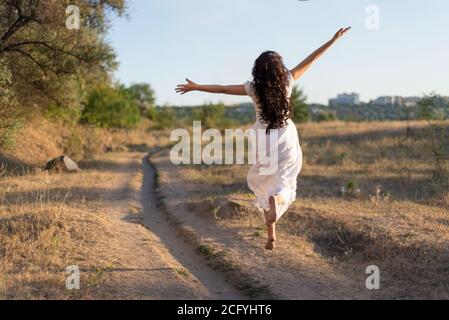 une fille heureuse court dans le champ le long du chemin, rebondissant et agitant ses bras. Sur fond de soleil d'été. Banque D'Images