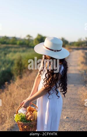 jeune fille dans une robe et un chapeau blancs, tenant un panier de fruits dans ses mains, sur le fond du soleil couchant l'été. Banque D'Images