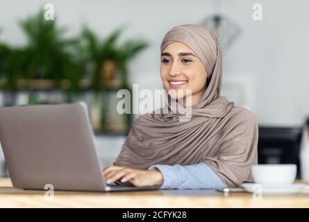 Joyeuse fille musulmane dans hijab travaillant en ligne au café Banque D'Images