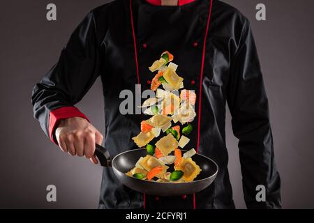 Gros plan du chef qui jette des pâtes italiennes avec des légumes et de la salmone dans l'air. Concept de préparation des aliments Banque D'Images