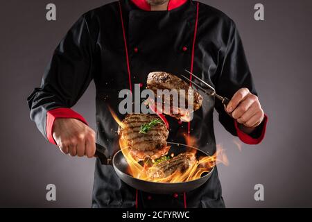 Gros plan du chef qui jette des steaks de bœuf dans l'air, feu de flammes autour. Concept de préparation des aliments Banque D'Images
