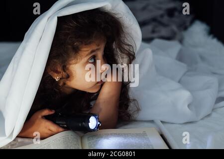 Une petite fille lit un livre intéressant avec une lampe de poche sous une couverture Banque D'Images