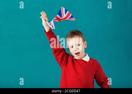 Vacances scolaires en Grande-Bretagne. Petit écolier avec le drapeau national du Royaume-Uni ayant du plaisir contre le mur bleu. Anglais pour enfants con Banque D'Images