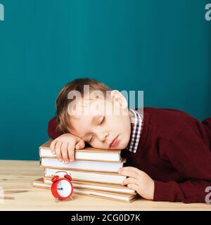 un garçon d'école fatigué dormait sur des livres. un petit étudiant dormant sur des manuels scolaires. L'enfant en uniforme scolaire se trouve sur la table avec une grosse pile de livres contre le bleu Banque D'Images