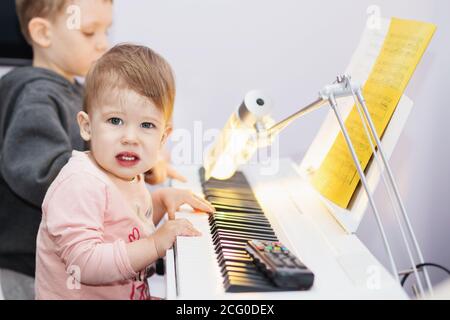 La petite fille aime jouer du piano électrique (synthétiseur) pour la première fois. Les petits enfants jouent le synthétiseur de piano à la maison avec Piano Score Sheet Music Banque D'Images