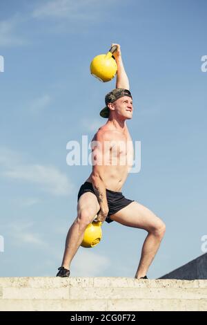 Portrait en coupe basse d'un homme méconnaissable qui soulève deux lourdes cloches de kettlebell avec un effort vigoureux pendant l'entraînement à la salle de gym Banque D'Images