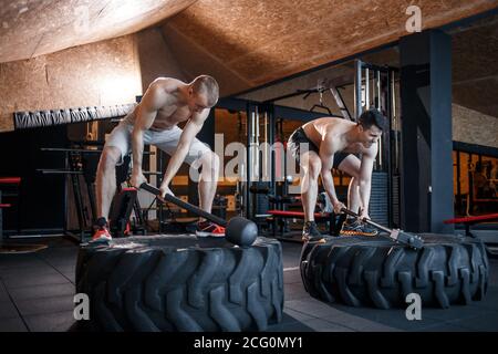 Deux jeunes hommes avec un marteau frappe sur un pneu dans la salle de gym. Entraînement. Banque D'Images