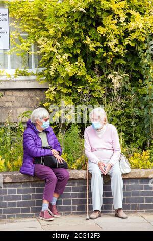 Deux femmes âgées retraités portent des masques sociaux distancé assis sur un mur parlant pendant le virus corona pandémie covid 19 Banque D'Images