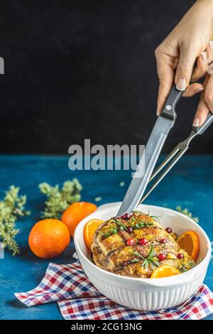 Femme en train de préparer de savoureux porc rôti dans un plat blanc, jambon cuit de noël aux canneberges, mandarines, thym, romarin, ail sur table légère sur Banque D'Images