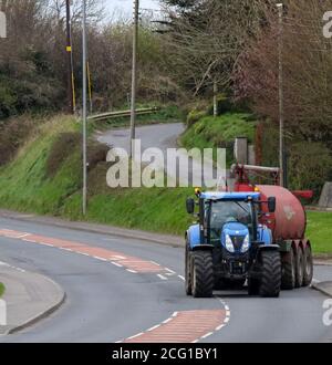Tracteur New Holland tirant un camion-citerne à lisier de ferme rouge entrant dans une route principale à partir d'une route rurale au printemps. Banque D'Images