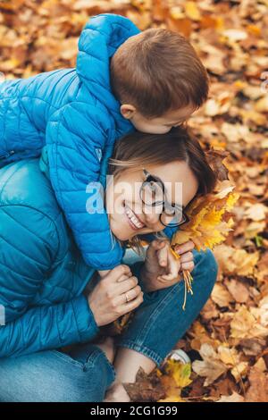 Vue de dessus photo d'une mère et d'un fils caucasiens dedans vêtements bleus jouant sur le sol avec des feuilles pendant un promenade automnale dans le parc Banque D'Images