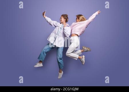 Deux charmantes femmes en vêtements lumineux et cheveux blonds sautant sur un mur de studio violet et de faire un selfie en utilisant un téléphone Banque D'Images