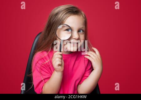 Jolie fille blonde utilisant un verre de loupe tout en recherchant quelque chose et sourire sur un mur rouge de studio dans le fauteuil Banque D'Images