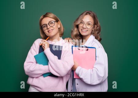 Photo en gros plan de deux sœurs blondes avec des lunettes souriantes à la caméra posé sur un mur de studio vert avec certains dossiers scolaires Banque D'Images