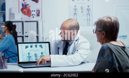 Jeune médecin masculin expliquant comment éviter les problèmes d'os à la vieille femme âgée à la retraite. Radiologie et radiographie dans un hôpital ou une clinique privé moderne avec le personnel médical marchant en arrière-plan, le personnel infirmier travaillant, le système de santé. Banque D'Images