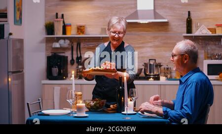 Vieille femme de haut niveau servant son mari avec du raisin et du fromage. Vieux couple parlant, assis à la table dans la cuisine, appréciant le repas, célébrant leur anniversaire à la maison avec de la nourriture saine. Banque D'Images