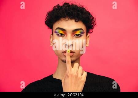 Gay homme portant l'ombre des yeux de couleur arc-en-ciel et smiley visage fingnail mettant son doigt dans les lèvres, gesturant pour être tout à fait. Homme transgenre gestante si Banque D'Images