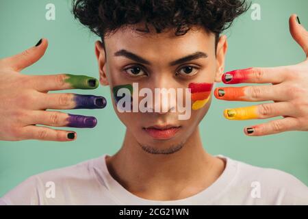 Gros plan d'un homme aux couleurs du drapeau LGTB peint sur son visage par deux mains. Homme gay avec drapeau de fierté peint sur ses joues. Banque D'Images