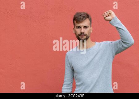 Un homme déterminé qui se relève le poing dans l'air sur fond de mur rouge. Jeune homme démontrant le pouvoir. Concept de protestation. Banque D'Images